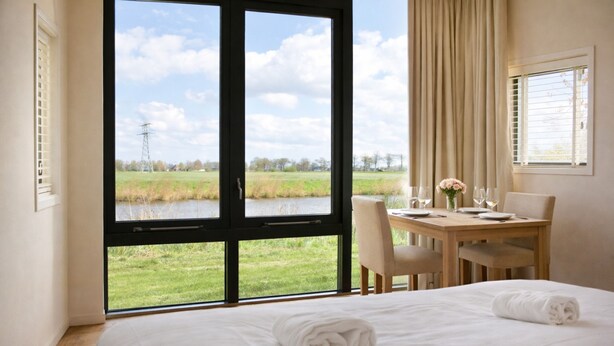 Une chambre d ’hôtel avec une grande fenêtre, un lit avec deux serviettes, une table à manger avec des chaises, et une vue sur un champ verdoyant et une ligne électrique.