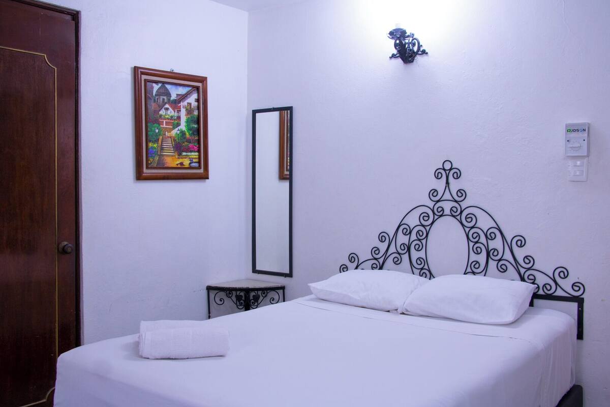 A bedroom with a bed, a wooden door, a painting, a mirror, and a wall-mounted light fixture.