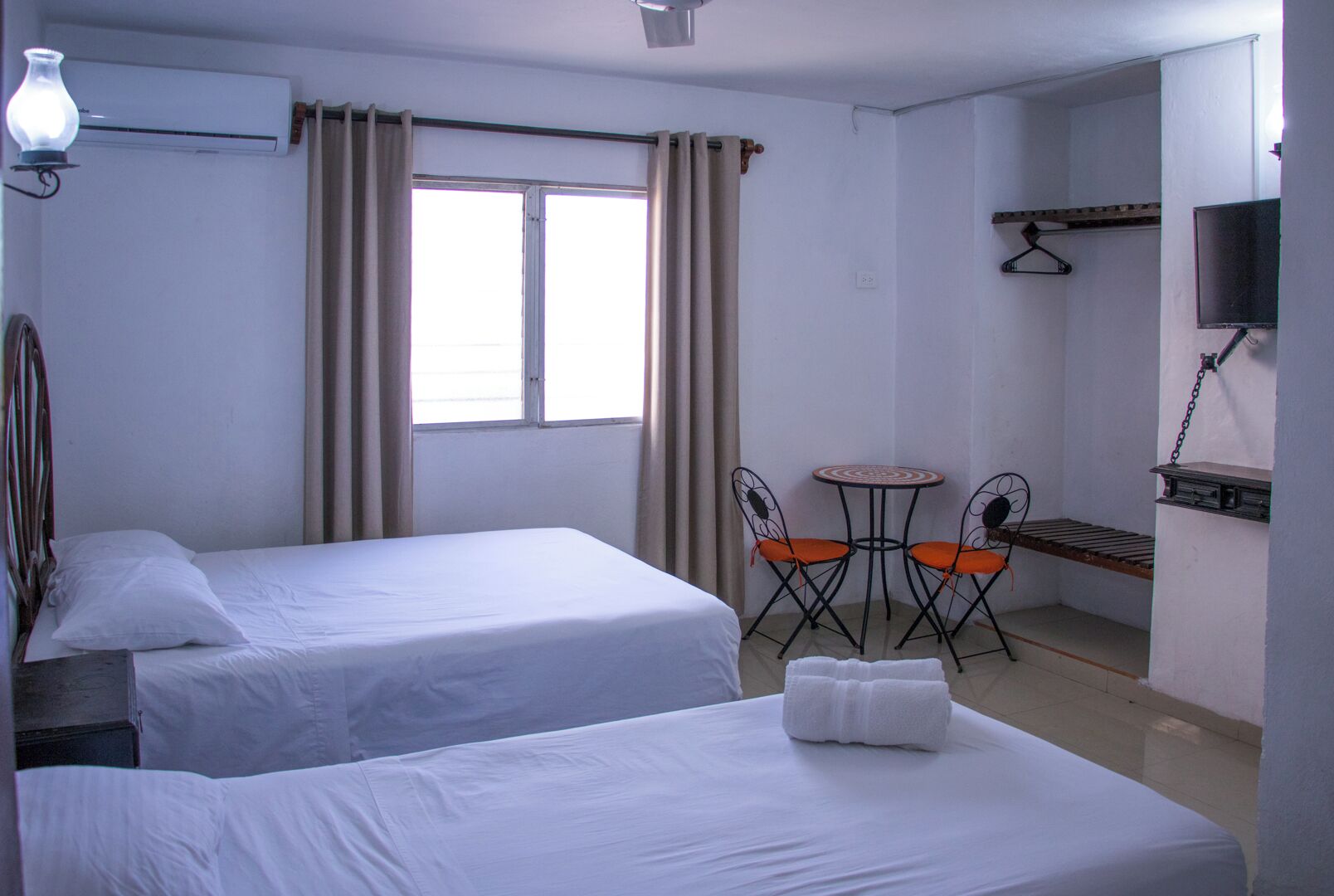 Habitación de hotel con dos camas, una pequeña mesa con sillas, un televisor montado en la pared y una ventana con cortinas.