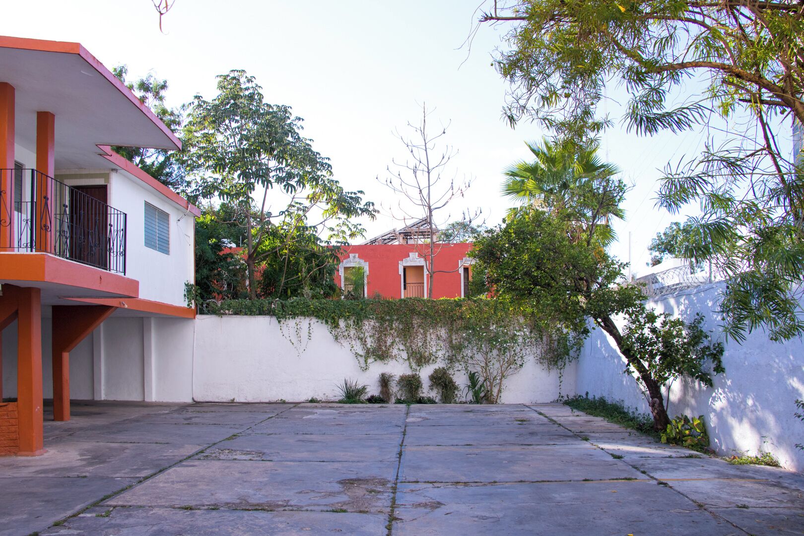 A courtyard with a white wall, a red building in the background, and a balcony with a railing.
