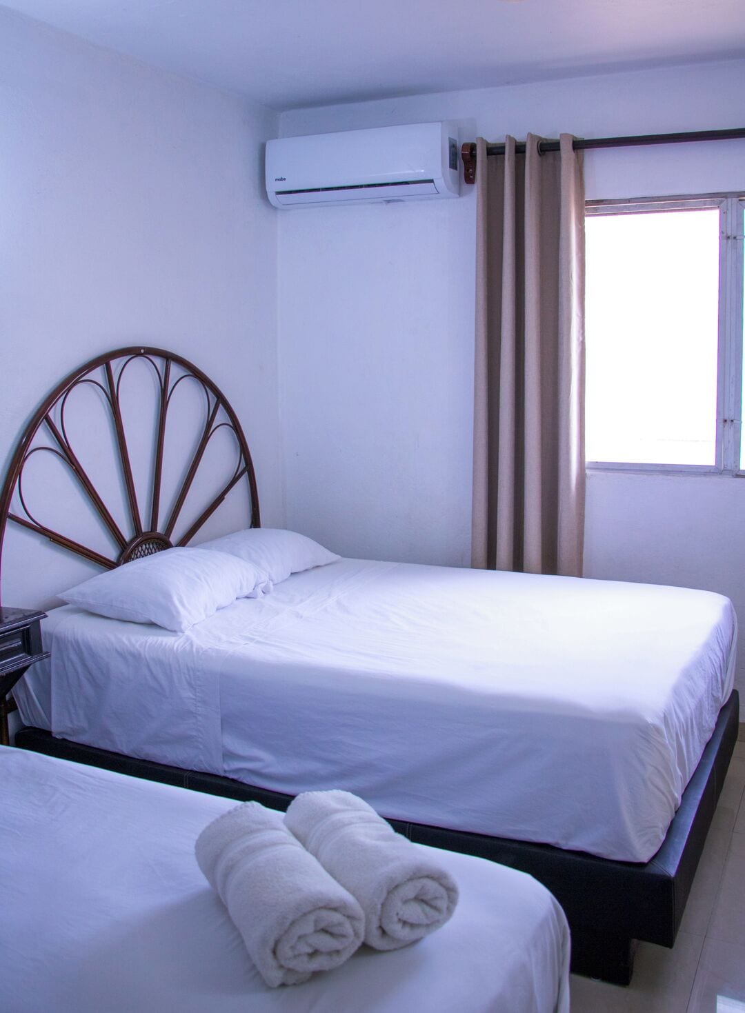 A hotel room with two beds, a wooden headboard, white bedding, an air conditioning unit, and a window with curtains.