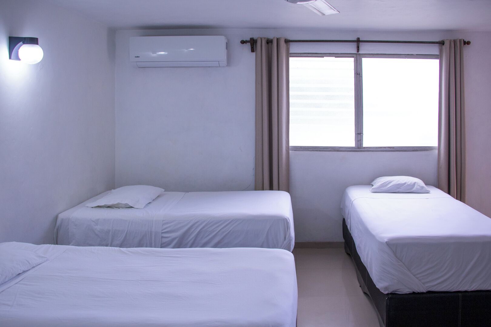Une chambre avec deux lits simples, un ventilateur de plafond, un appareil de climatisation, une fenêtre avec des rideaux et un luminaire fixé au mur.