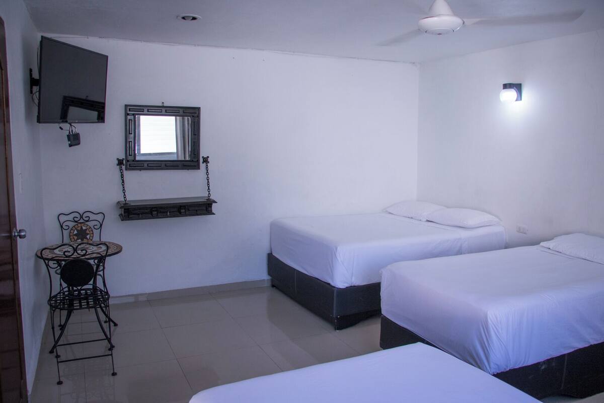 A room with two beds, a small table with a chair, a TV mounted on the wall, and a ceiling fan.