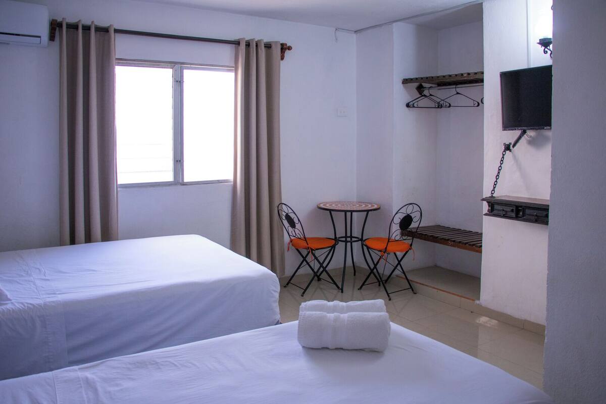 A hotel room with two beds, a small table with chairs, a TV, and a window with curtains.