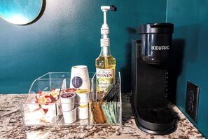A kitchen counter with a coffee maker, a bottle of Monin vanilla syrup, and a clear organizer containing various condiments and utensils. - Hollyhock Suite 411: 1 bed/1 bath (Louisville)