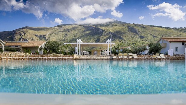 Una piscina amb un paisatge de muntanya al fons, una zona de seients coberta i una paret de pedra.