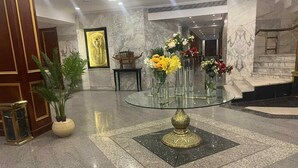 Ένας χώρος υποδοχής με ένα τραπέζι με γυάλινη επιφάνεια, το οποίο περιβάλλεται από διακοσμητικά βάζα με λουλούδια, μαρμάρινοι τοίχοι και μια κορνιζαρισμένη εικόνα που απεικονίζει ένα αιγυπτιακό αντικείμενο τέχνης.