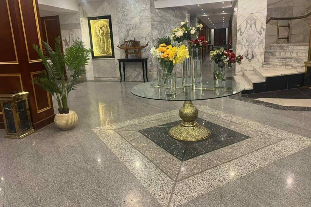 Un hall d’entrée avec une table à plateau de verre, entourée de vases de fleurs, des murs en marbre et un tableau encadré représentant un artefact égyptien.