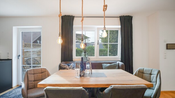 A dining area with a wooden table, four upholstered chairs, a vase with a bottle, and a window with curtains. - Allgäu 17 – Modern Alpine-Style Apartment (Leutkirch im Allgäu)