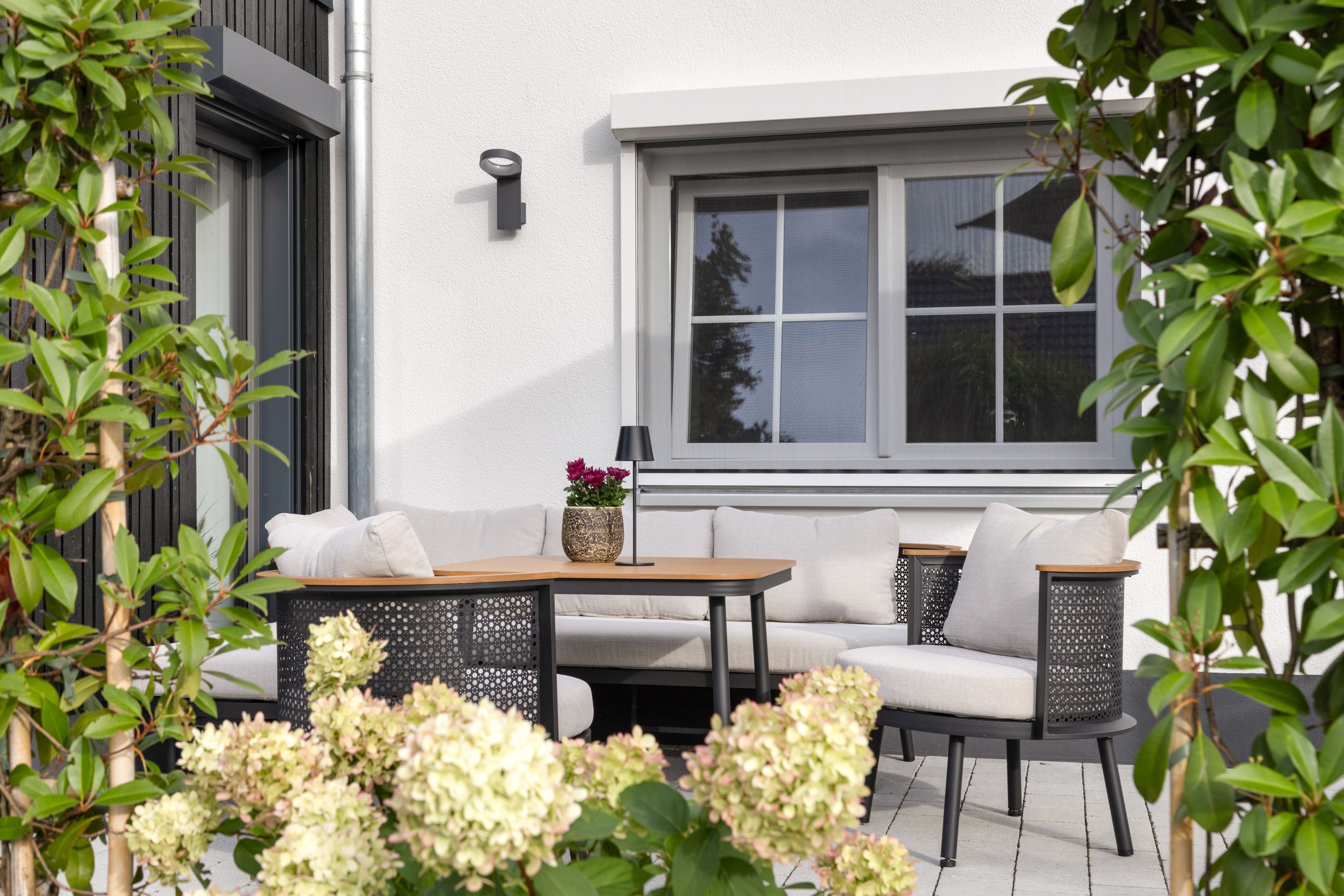 An outdoor seating area with a table and two chairs, surrounded by greenery and a white building with a window.