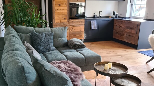 A modern living room with a grey sofa, a small round table with candles, and a kitchen area with wooden cabinets and a window. - Allgäu 17 – Modern Alpine-Style Apartment (Leutkirch im Allgäu)