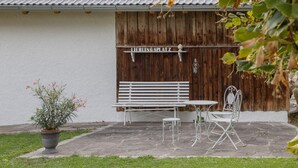 A patio with a white bench, a metal table, and chairs, a potted plant, and a wooden fence with a sign. - Allgäu 17 – Modern Alpine-Style Apartment (Leutkirch im Allgäu)