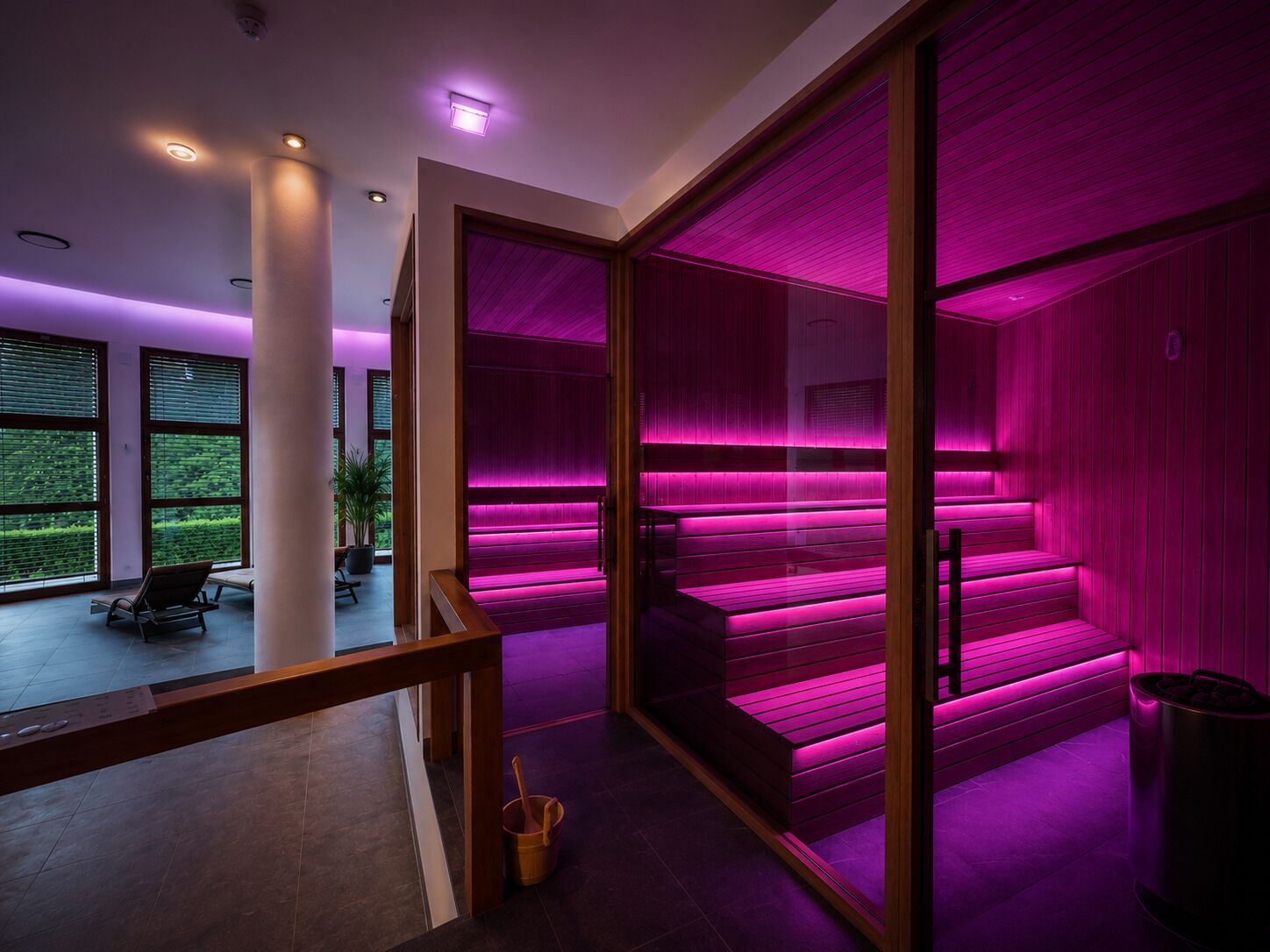 Un interior moderno con una escalera iluminada con luz púrpura, ventanas amplias y vistas al verde exterior.