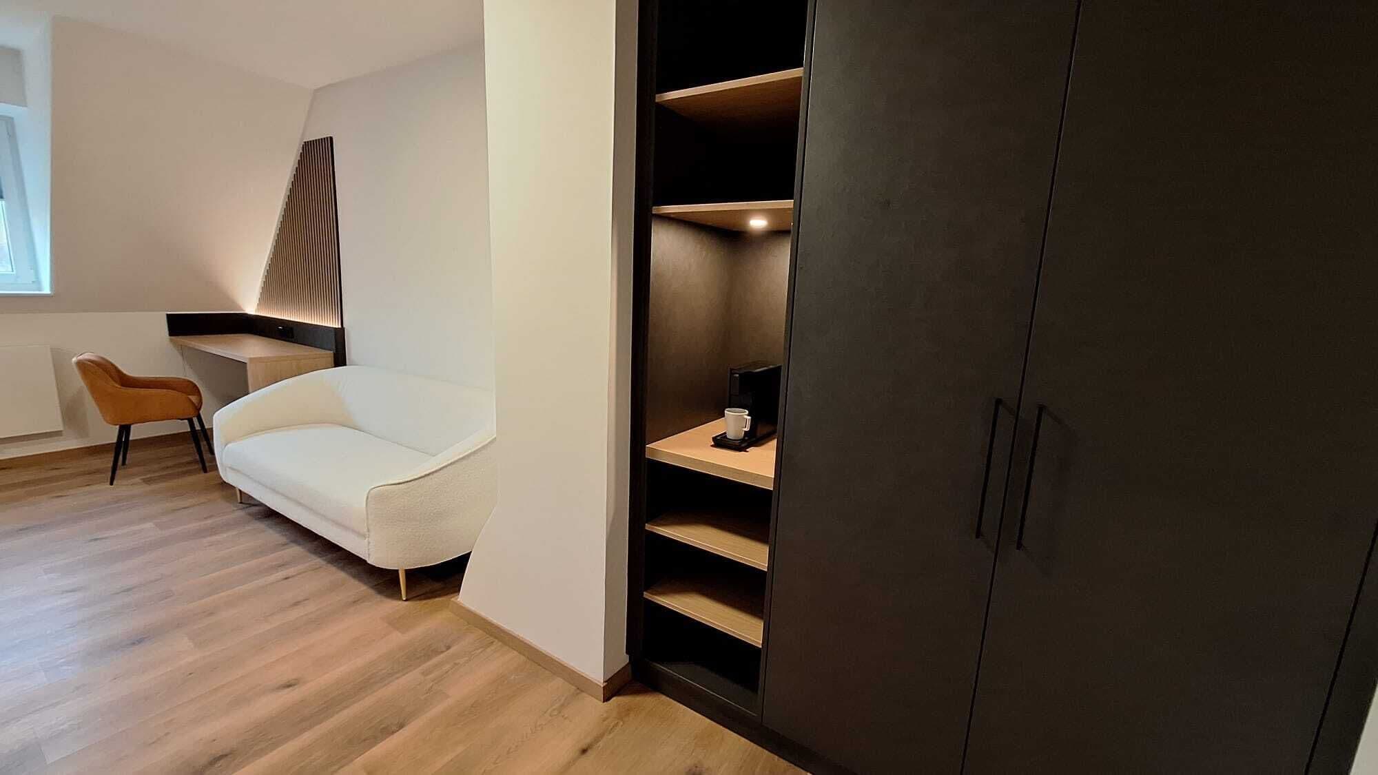 Une pièce moderne avec un canapé blanc, une chaise en bois, un meuble sombre et une étagère intégrée sur laquelle se trouve une machine à café.