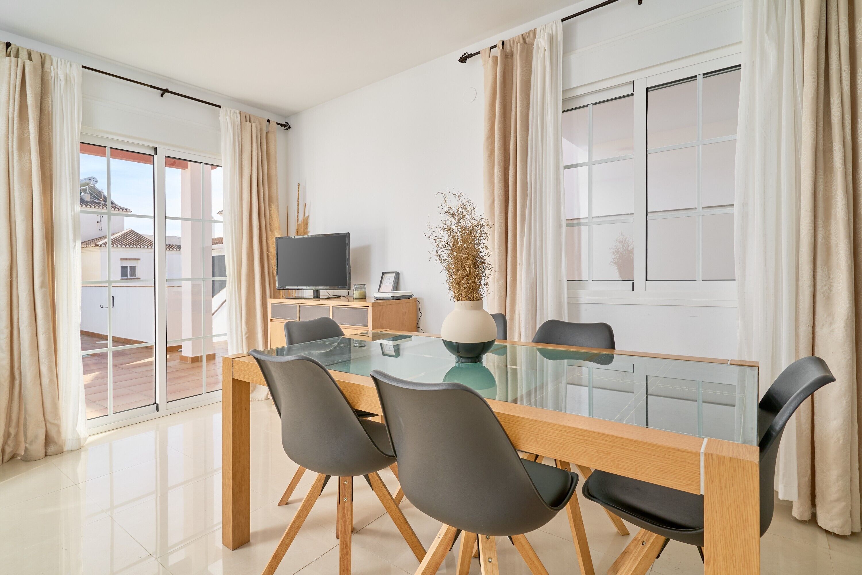A modern dining area with a glass table, four chairs, a television, and a vase with dried plants.