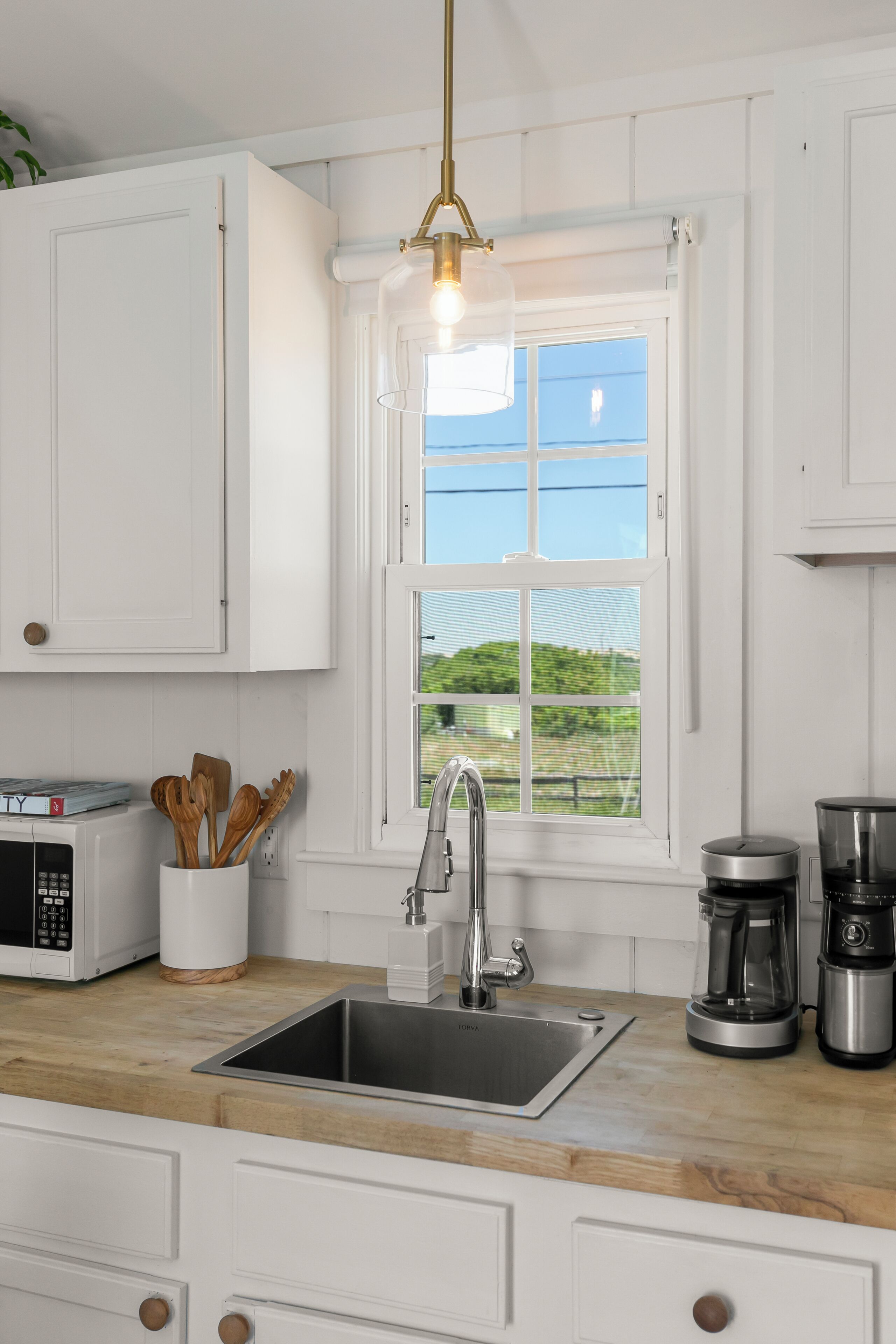 A kitchen with white cabinets, a wooden countertop, a stainless steel sink, a coffee maker, and a window with a view of greenery.