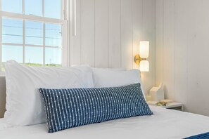 A bed with white and blue patterned pillows, a bedside table with a lamp, and a window with a view. - Private Beachfront Cottage for 2—Spacious Deck on the Water, Breathtaking Views! (Truro)