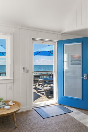 A room with a view of the ocean, a blue door, a wooden table with books, and a mat. - Private Beachfront Cottage for 2—Spacious Deck on the Water, Breathtaking Views! (Truro)