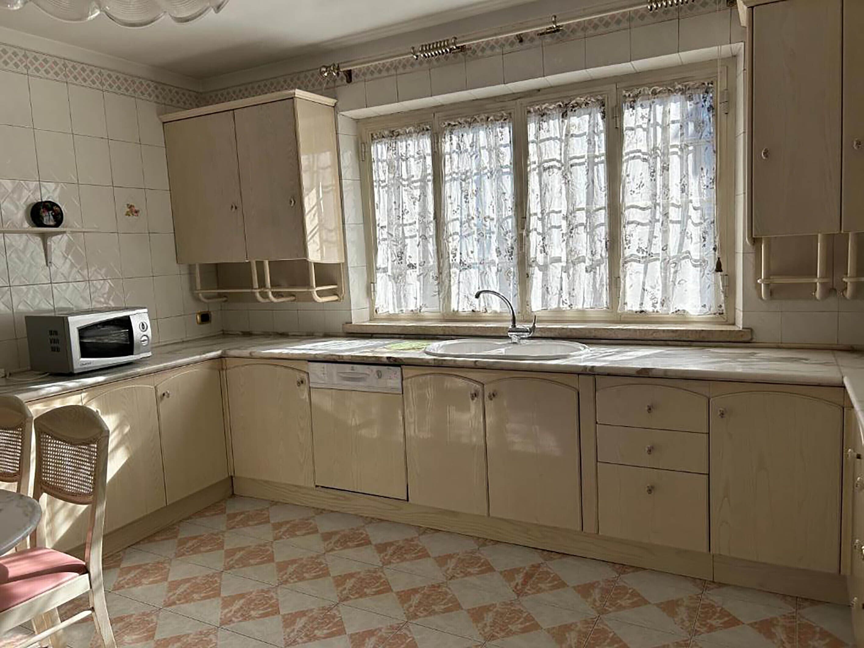 A kitchen with a microwave, sink, and window with lace curtains.