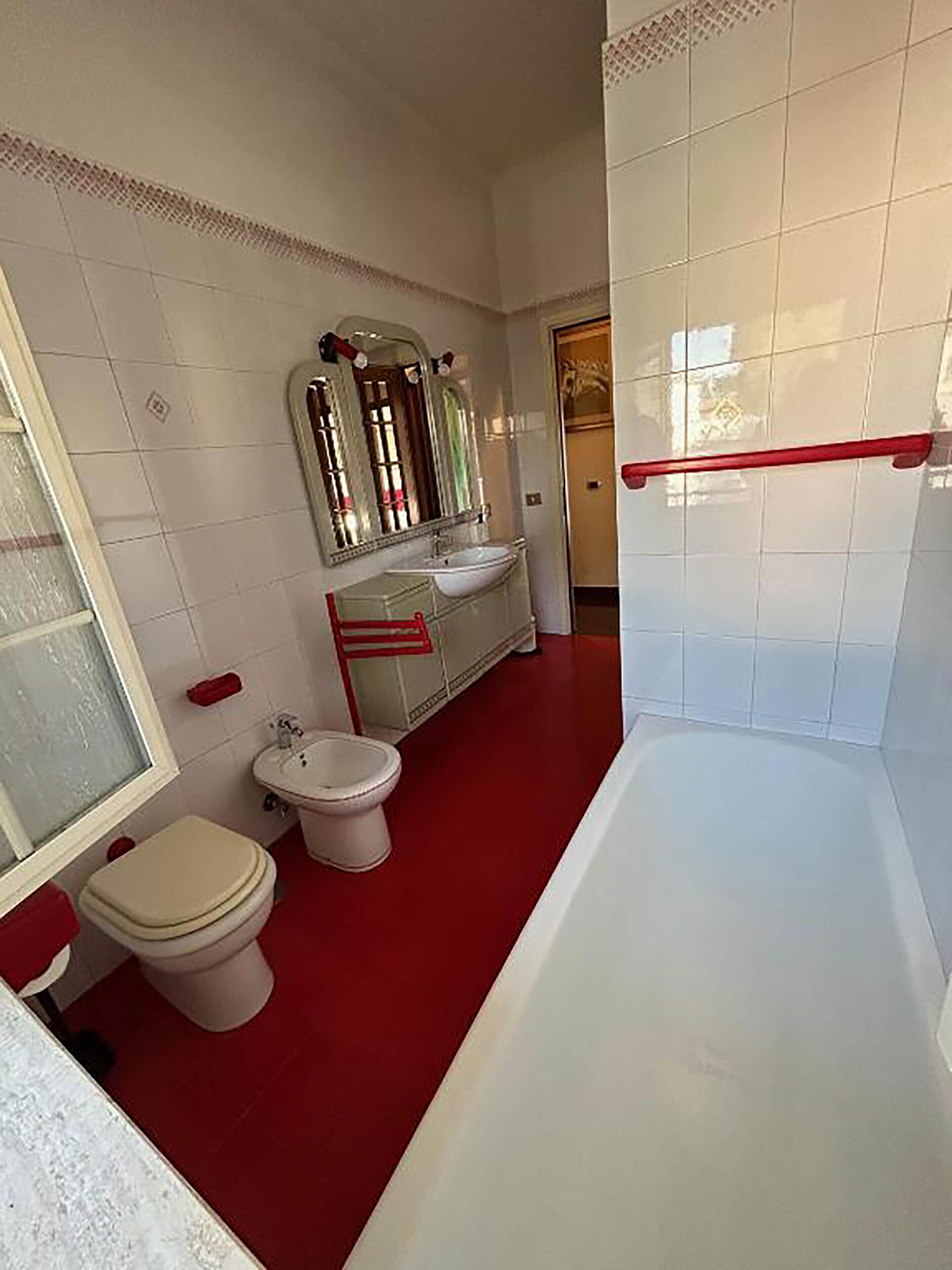 Ein Badezimmer mit rotem Boden, weißen Armaturen, einem großen Spiegel und einem roten Handtuchhalter.