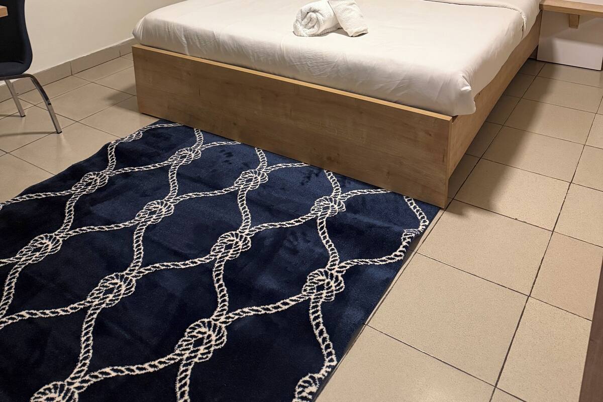 Ein Schlafzimmer mit Bett, Nachttisch, Sessel, Fenster mit Blick auf Gebäude und einem Bild an der Wand.