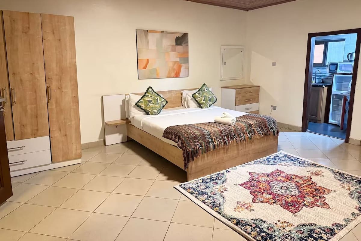 Ein Schlafzimmer mit Bett, Nachttischen, einem Holzschrank, einem großen Fenster und einer durchgehenden Tür erkennbaren Küche.