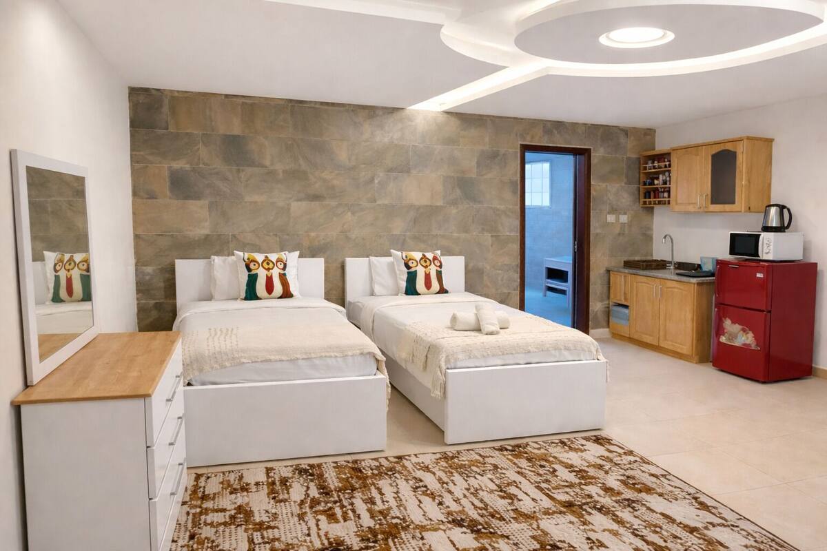 Ein modernes Hotelzimmer mit zwei Betten, einer Holzkommode, einer Küchenzeile inklusive rotem Kühlschrank und einer Wand aus Stein.