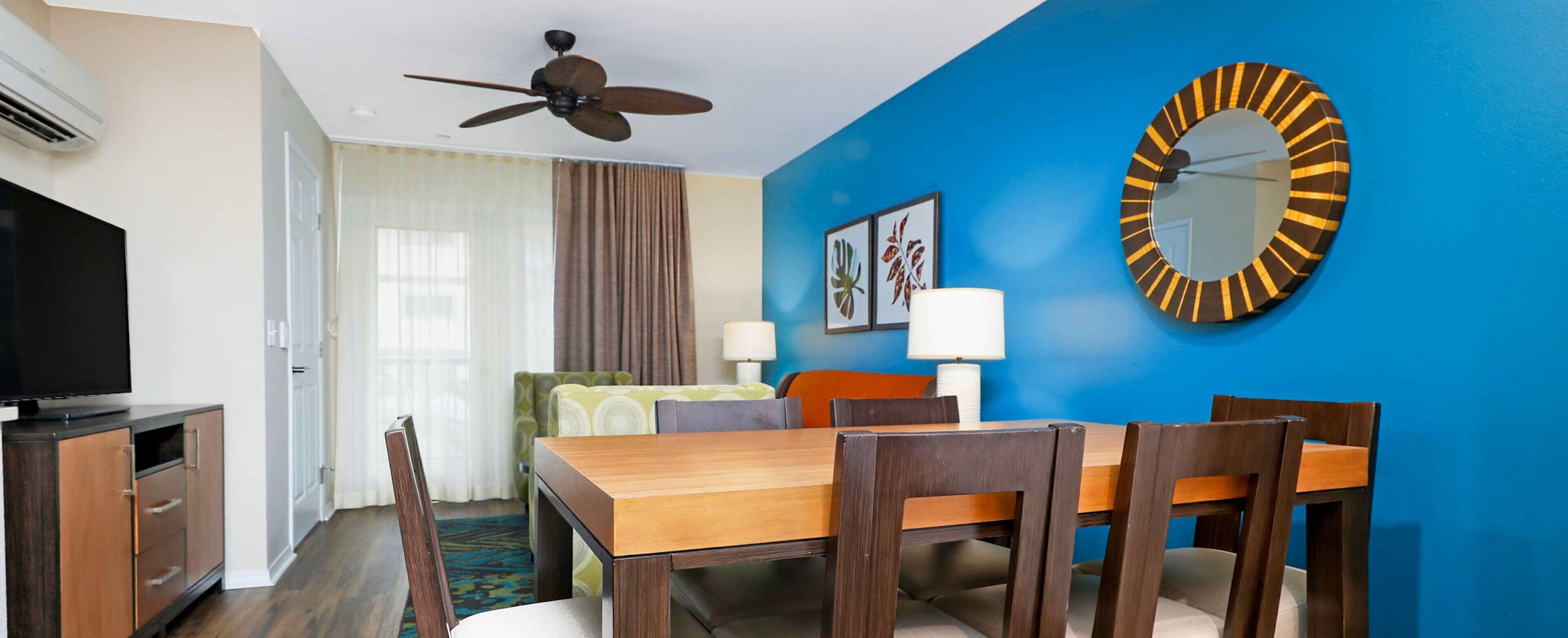 A hotel room with a dining area, a ceiling fan, a flat-screen TV, and a large mirror.