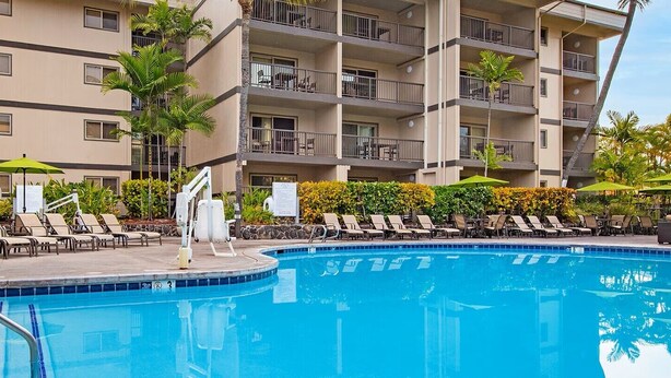 A swimming pool with lounge chairs and umbrellas, surrounded by a multi-story building with balconies.