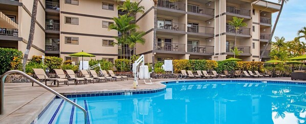 A swimming pool with lounge chairs and umbrellas, surrounded by a multi-story building with balconies.