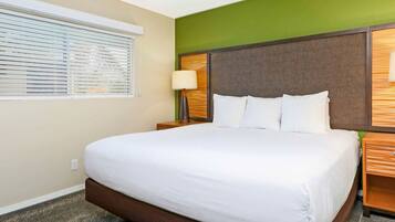 A hotel room with a large bed, two bedside tables, a nightstand, a window with blinds, and a green accent wall.