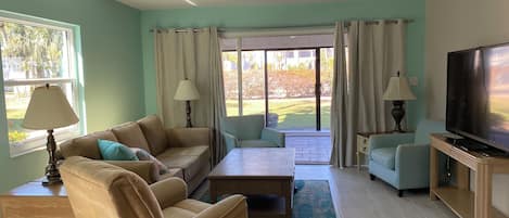 A living room with a sofa, armchair, coffee table, and TV. There are two lamps, a window with curtains, and a view of a garden.