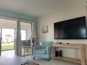 Ein Wohnzimmer mit blauem Ohrensessel, einem Fernseher auf einem Schrank, einem kleinen Tisch mit Lampe und Blick ins Freie durch eine Glastür.
