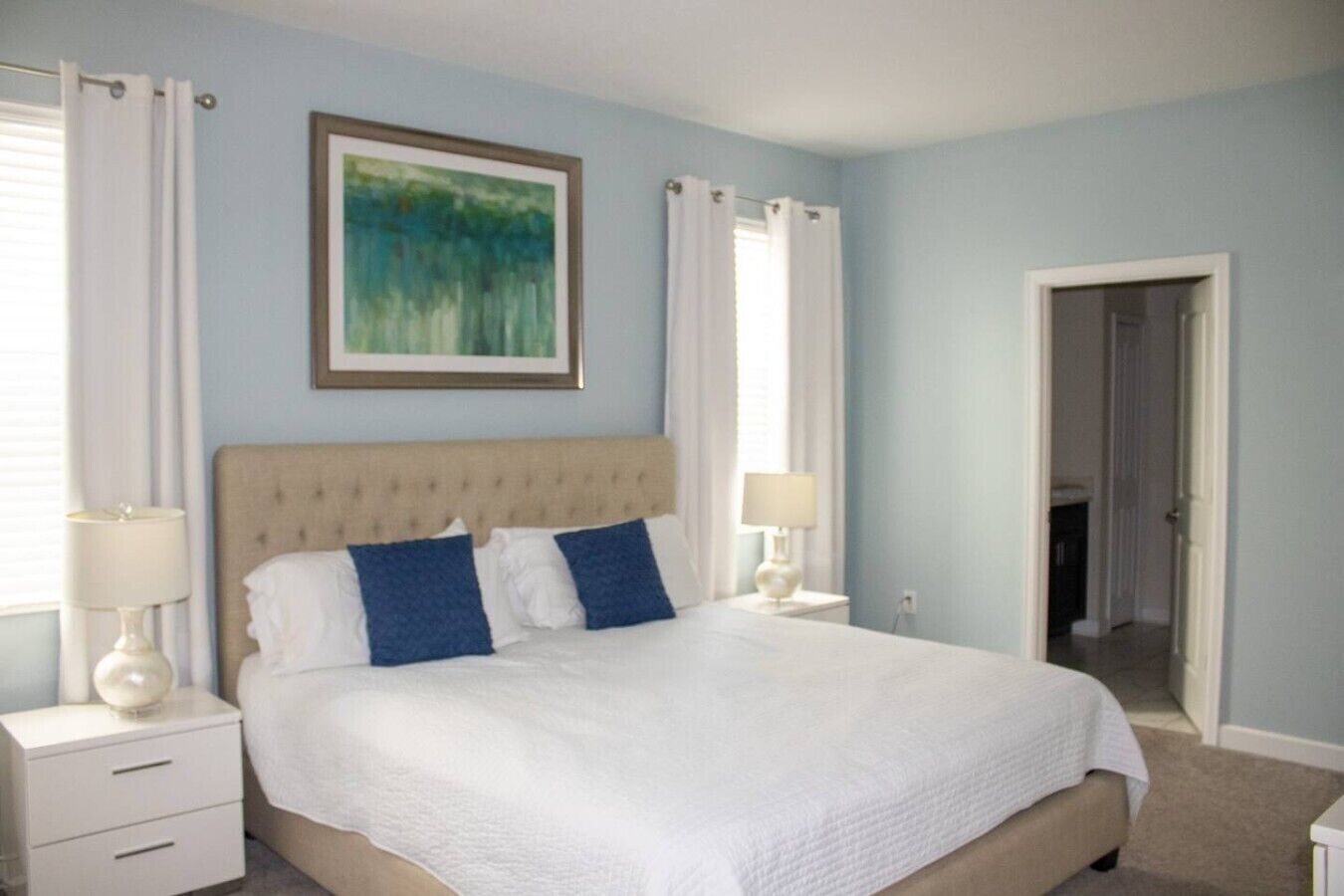 A bedroom with a bed, two bedside tables, a nightstand, a painting, and a door leading to another room.