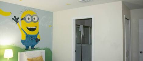 A bedroom with a bed, a nightstand, a wall mural, a door, and a ceiling light fixture.