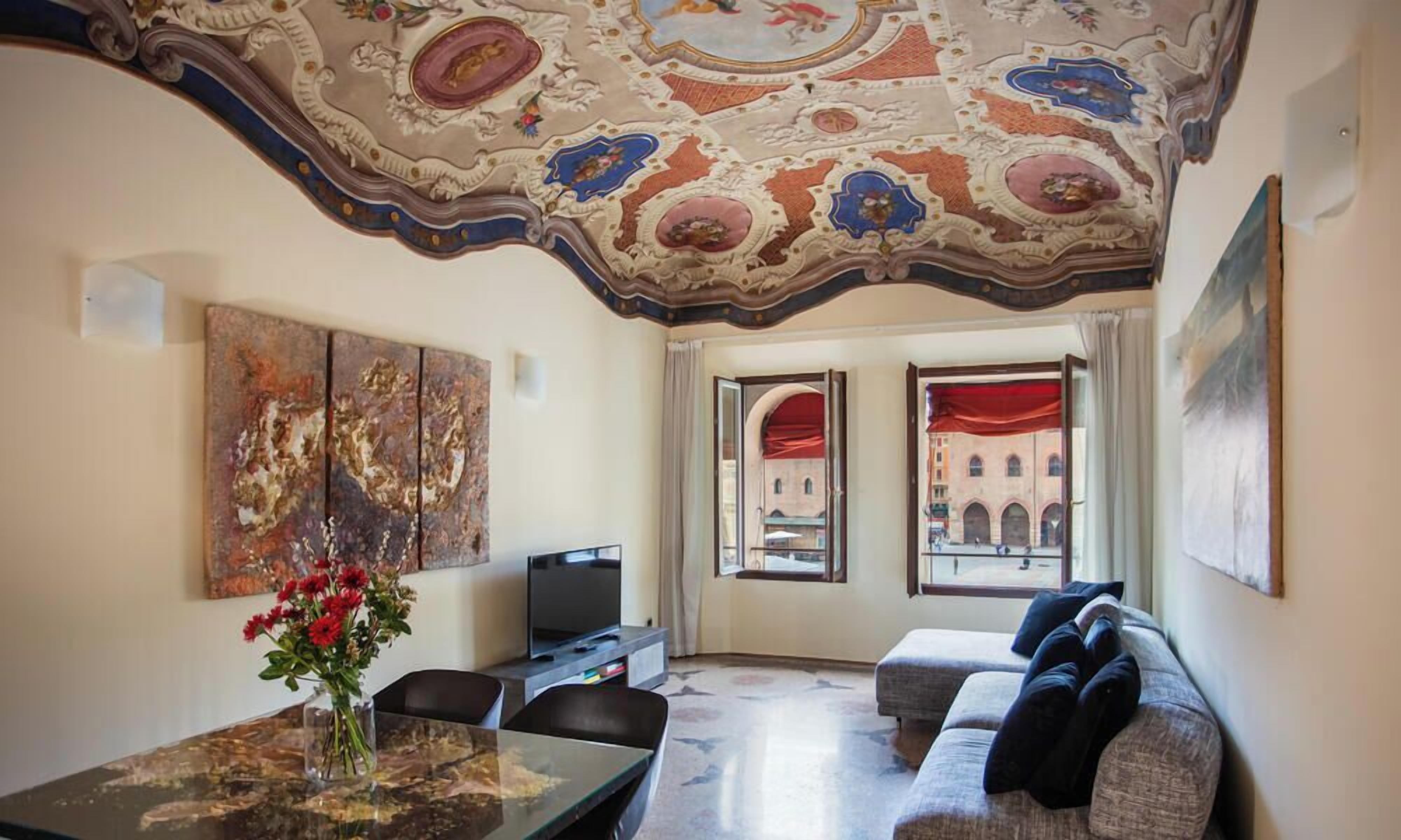 Una sala de estar con una mesa de vidrio, un sofá y un televisor. Se ve una ventana con vistas a edificios.