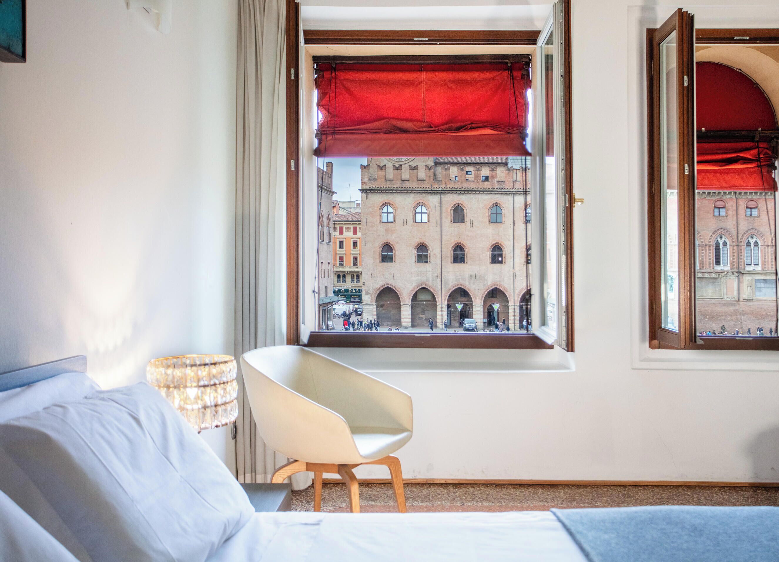 Ein Hotelzimmer mit Bett, Sessel, Fenster mit rotem Rollo und Blick auf ein historisches Gebäude.