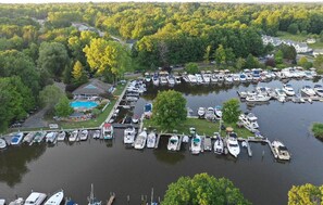 Vista aérea de uma marina com diversos barcos atracados, cercada por vegetação densa e um pequeno edifício com piscina.