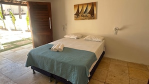 Un letto rifatto con una coperta color petrolio e cuscini bianchi, un quadro con dei velieri sulla parete e una porta in legno che dà su un'area esterna.