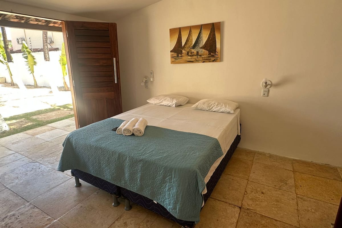 Een net opgemaakt bed met een blauwgroene spreidek en witte kussens, een schilderij van zeilboten aan de muur, en een houten deur die leidt naar een buitenruimte.