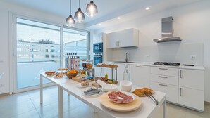 A modern kitchen with a large island, white cabinetry, and a view through a glass door. - Aura B&B - Queen Room with Jacuzzi and Private Terrace (Pachino)