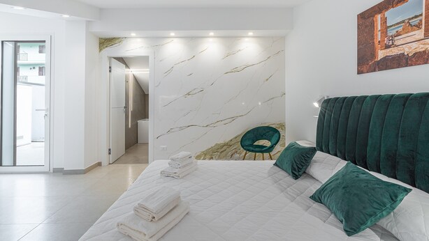 A bedroom with a bed, green pillows, a green chair, a marble-patterned wall, and a painting. - Aura B&B - Queen Room with Jacuzzi and Private Terrace (Pachino)