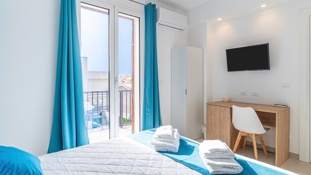 A hotel room with a bed, a desk, a TV, and a balcony with curtains. - Aura B&B - Quadruple Room Bed and Breakfast (Pachino)