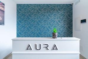 Un mostrador de recepción con señalización 'AURA', una pared de diseño azulado y una pequeña estatua de Buda.