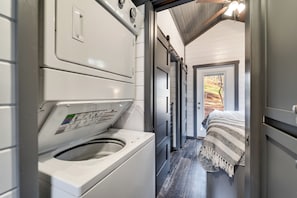 Une buanderie équipée d’un lave-linge, d’un sèche-linge et d’une porte donnant sur une chambre avec un lit et une vue sur les arbres.