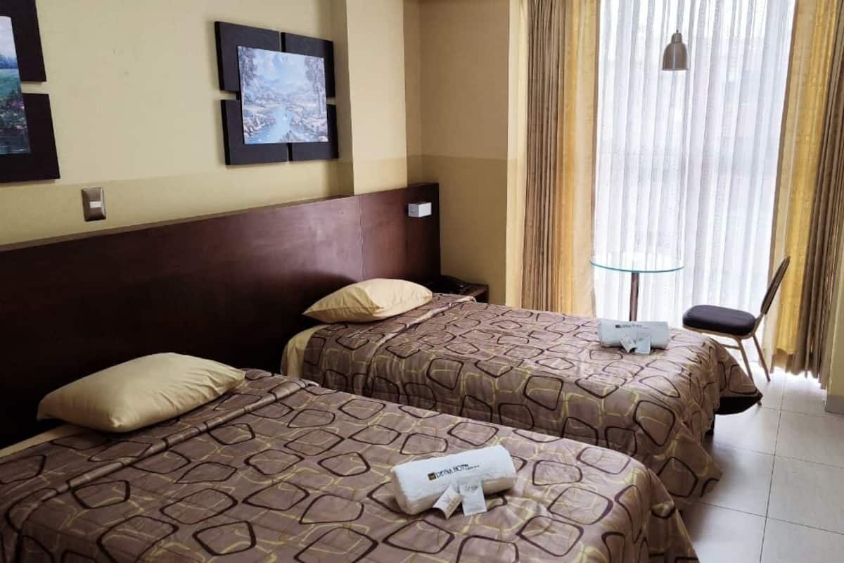 A hotel room with two beds, a small table, a chair, and a window with curtains.