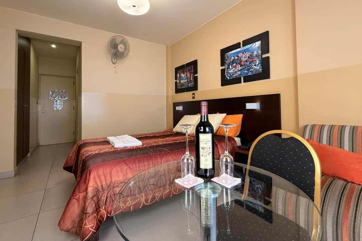 A hotel room with a bed, a glass table with a bottle and glasses, a TV, and a wall-mounted fan.
