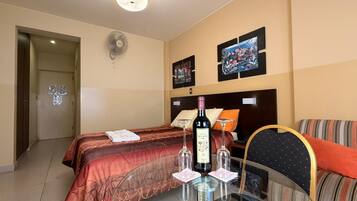 Habitación de hotel con cama, una mesita de vidrio con una botella y vasos, televisor y ventilador de pared.