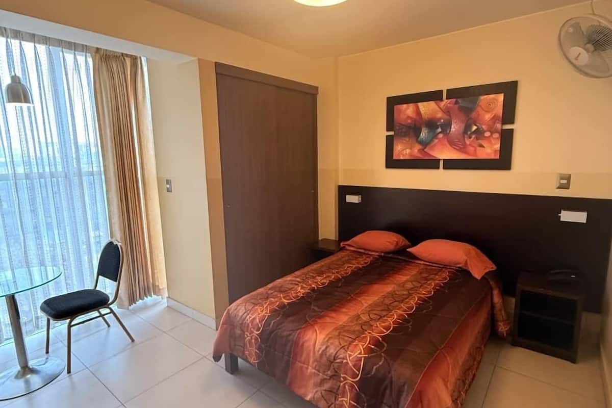 A hotel room with a bed, a chair, a desk, a window with curtains, a ceiling light, a wall-mounted fan, and a painting.