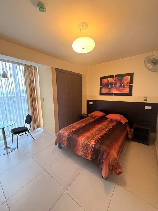 Une chambre d’hôtel comprenant un lit, une chaise, un bureau, une fenêtre avec des rideaux, un luminaire au plafond, un ventilateur fixé au mur et un tableau.